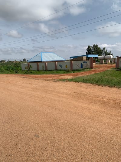 The image depicts an outdoor scene in Nabatum, Nigeria. In the foreground, a dirt road takes up a significant portion of the frame, with patches of green grass growing along its edges. Behind the road, a low, light-colored wall runs horizontally across the mid-ground. The wall is adorned with simple, colorful painted images, including what appear to be bottles and a gas cylinder. A blue and white building with a peaked roof is visible behind the wall on the left side of the image. To the right of this building, a more open area reveals a large white cylindrical tank, likely a storage tank, elevated on supports. A person is seen walking near this tank. In the background, lush green vegetation and trees are present, under a partly cloudy sky. Power lines are strung across the upper portion of the image.