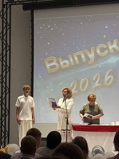 In Markovskii, Russia, on a brightly lit stage, a young man in a white polo shirt and pants stands to the left, facing forward. To his right, a woman with curly blonde hair, dressed in a white suit, speaks into a microphone, holding a document. Behind her, another woman in a green shirt and glasses stands behind a table covered with a red tablecloth, organizing papers. The backdrop features a projected image with the text "Выпуск 2026" (Graduation 2026) in large gold letters. Several people are visible in the audience in the foreground, some with balloons, suggesting a graduation ceremony.