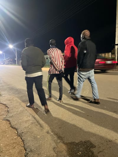 Four individuals are walking away from the camera on a paved road at night in Barakin Lerawi, Nigeria. The road appears to be an unlit street, possibly a residential or less developed area, with a cracked and uneven surface. Streetlights are visible in the background, casting long shadows of the people and creating a dimly lit atmosphere.
The person closest to the camera on the left is wearing a dark long-sleeved shirt and black pants, with sandals on their feet. To their right, another person is wearing a red hooded jacket and dark pants, also with sandals. Next to them, a person in a vertically striped shirt (red and white) and dark pants with yellow stripes is walking. The furthest person on the right is wearing a black jacket, light blue jeans, and sandals. The group appears to be casually strolling. In the background, a red car with illuminated taillights is partially visible, suggesting traffic or parked vehicles. Buildings and what might be wooden structures are present further in the background, partially obscured by darkness and distance. The overall scene depicts a nighttime street environment in a Nigerian locality.