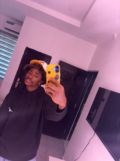 A person with dark skin and short, curly hair, wearing a yellow and white baseball cap and a black hoodie with a logo visible on the lower right side, is taking a selfie indoors. The phone in their hand has a yellow case. Behind them, there's a dark-colored door and a sleek, dark-colored object mounted on the wall near the ceiling, potentially a TV or a decorative panel. The walls are a light shade, and there is a ventilation unit visible on the lower left. The scene seems to be in Igboefon, Nigeria.