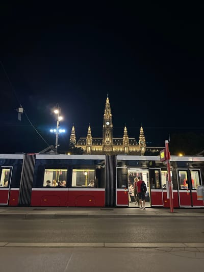 The image shows a night scene in Vienna, Austria, with the Rathaus (City Hall) illuminated in the background. A red and black tram with several passengers inside is stopped at a tram stop. Two people are standing at the open doors of the tram, one appearing to be boarding and the other waiting. The street in front of the tram is paved, and a sidewalk runs alongside it. The sky is dark and clear, indicating nighttime.