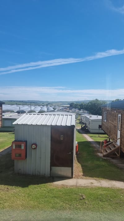 This media file shows a row of prefabricated buildings or cabins, likely in a rural setting given the presence of green fields and distant wind turbines. The buildings appear to be constructed of metal and wood, with some having brown doors and windows. In the foreground, a silver building with a white corrugated metal roof is prominent. It has a brown door and a red fire alarm mounted on its side, as well as a smaller orange box below it. The number '2' is visible on the brown door.

Behind this building, more similar structures are visible, stretching into the distance. The sky is a clear, bright blue with prominent white contrails, suggesting it is daytime. The ground is covered in grass, with a paved path running alongside the foreground building. The overall scene suggests an industrial or perhaps a temporary accommodation site in a well-lit, clear weather environment. There is no activity or people visible in the frame. The location context provided is Auchenblae, United Kingdom.