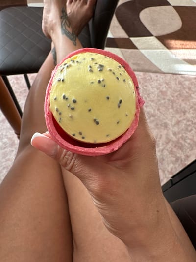 A close-up, overhead shot shows a person's hand holding a bright pink, waffle-like cone filled with a round scoop of yellow ice cream studded with black seeds, likely chia seeds. The ice cream is a light yellow, and the cone has a textured, crisp appearance. The person's hand, with manicured nails, is holding the cone from the side, obscuring some of the cone's base. The background is blurred, revealing glimpses of a person's legs and feet, wearing visible tattoos on their ankles and feet. The legs are crossed, suggesting a seated or relaxed posture. A dark chair and a patterned rug with brown and beige squares are partially visible. The overall lighting suggests an indoor setting, possibly a home. The city of Yuzhnouralsk, Russia, is not depicted in this image.
