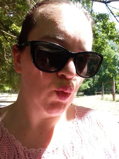 A close-up, outdoor, selfie-style photograph shows a woman wearing sunglasses and a pink crocheted top. She is outside, with trees and a fence visible in the background. The woman is puckering her lips. The image is taken from a low angle, looking up at the woman's face. The lighting suggests it is daytime.