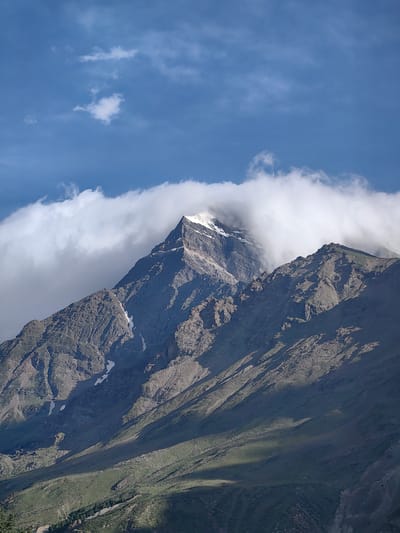 A vertical, medium shot captures a majestic mountain peak in Yamling, India, partially obscured by a dramatic band of white clouds that hugs its summit. The mountain itself is a study in rugged texture, with dark, rocky slopes marked by deep shadows that suggest steep inclines and rugged terrain. Patches of snow glint on the upper reaches of the mountain, hinting at its high altitude. The sky above is a vibrant blue, dotted with a few wispy white clouds. The lower slopes of the mountain transition into a softer, greener landscape, with visible vegetation and shadowed areas. The overall impression is one of immense natural beauty and the raw power of the mountains.