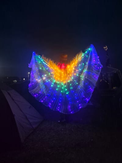 A person stands in an outdoor field at night, holding a large, fan-like costume adorned with colorful LED lights. The lights display a gradient of colors, including red, orange, yellow, green, and blue, radiating outwards from the center. The costume is translucent, allowing the lights to shine through. The person is wearing what appears to be a beaded headdress. In the background, several tents are visible, suggesting a camping or festival environment. The ground is covered in grass and possibly some scattered debris. The sky is dark and cloudless. The location is identified as Lagunovo, Russia.