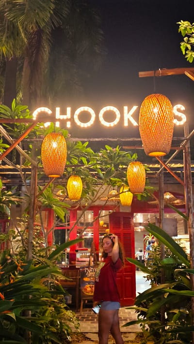 An outdoor night scene at what appears to be a restaurant or cafe in Nam Hoa Lư Ward, Vietnam, is captured. A neon sign spelling "CHOO KS" is prominently displayed overhead, illuminated by warm lights. Several rattan lanterns hang at various heights, casting a yellowish glow. Lush green foliage and palm trees surround the area, with a red building facade featuring shuttered windows visible in the background. A young woman with long, curly brown hair is standing in the foreground, partially obscured by the plants. She is wearing a maroon t-shirt and denim shorts, with sunglasses perched on her head. She is holding a smartphone in her right hand and touching her hair with her left. The ground is paved with stones. The overall atmosphere is tropical and inviting.