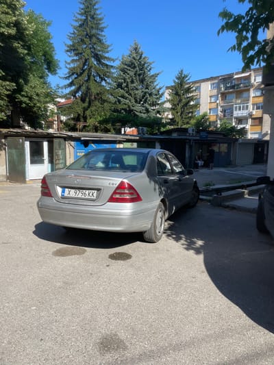 A silver Mercedes-Benz C-Class sedan is parked on an asphalt surface in front of a row of small garages. The car has a Bulgarian license plate that reads "X 9796 KK". The scene appears to be an outdoor urban environment, likely a residential area, with a multi-story apartment building visible in the background. Several tall evergreen trees are also present, casting shadows on the ground. The sky is clear and blue. In one of the garages, two individuals are visible: one person is seated, and another is standing, appearing to interact with the seated person. The overall lighting suggests it is a sunny day. The location is identified as Haskovo, Bulgaria.
