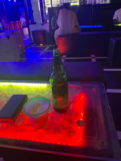 An empty green glass bottle lies horizontally on a table, alongside a translucent plastic cup filled with a white substance. A dark rectangular object, possibly a power bank, is near the cup. The scene is illuminated by red, yellow, and blue neon lights. The table appears to be made of glass or clear plastic, resting atop small white stones. In the background, a person is seated in a booth.