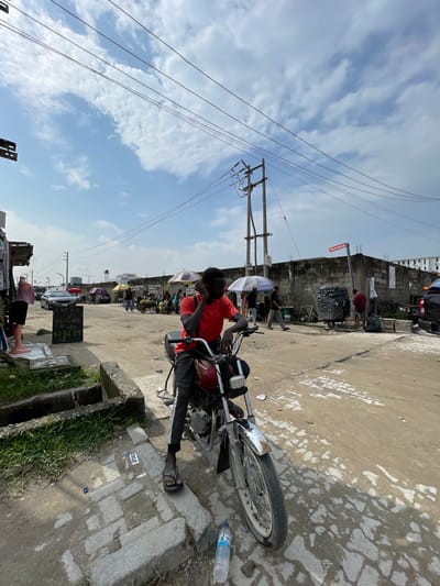 In Igboefon, Nigeria, a young man in a red t-shirt and dark pants sits on a red motorcycle, holding a phone to his ear. The scene is a dusty street with a partially paved road and patchy grass. In the background, a concrete wall, construction materials, and several people are visible, some holding umbrellas. There are also vehicles, including cars and trucks, and market stalls with produce and goods. The sky is blue with scattered clouds.