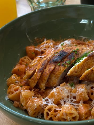 A close-up shot captures a bowl of pasta with sliced grilled chicken. The pasta appears to be a ridged, tube-shaped variety coated in a red sauce, possibly tomato-based, and topped with shredded white cheese and small green herbs, likely chives. The grilled chicken breast is sliced and seasoned, showing char marks on its surface. The dish is presented in a dark green ceramic bowl. In the blurred background, a glass of orange juice is visible to the left, and another out-of-focus glass is seen behind the bowl. The setting suggests a restaurant or dining environment. The image does not provide specific visual cues to pinpoint its location in San Pedro Garza García, Mexico.