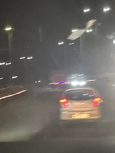 The image appears to be a photograph taken at night, possibly from inside a vehicle, given the motion blur and perspective. The scene is dark, with numerous lights that are blurred and streaky due to movement.
The primary subject appears to be a light-colored vehicle with its red taillights illuminated, seen from the rear. The vehicle is slightly tilted. In the foreground and scattered throughout the image are various bright, blurry lights, some appearing as streaks and others as more concentrated orbs. Some of these lights are white, while others have a reddish or yellowish hue, consistent with vehicle headlights or streetlights. There are also some indistinct shapes that might be a person or object, partially obscured and blurred. In the top left, there is a white, amorphous shape that is also blurred, possibly a piece of cloth or debris.
The overall impression is that of a moving scene at night, characterized by artificial lights and a lack of clear detail. There are no visible text elements in the image. The context provided, "B Camp, India," suggests the location, but this information cannot be visually verified from the image itself. The time of day is clearly nighttime, and the weather is not discernible. There are no discernible emotions conveyed due to the lack of clear subject detail and the abstract nature of the light streaks.