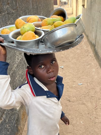 A young person with dark skin is carrying a metal container filled with mangoes, which rests on their head. The mangoes are yellow and green. The person is wearing a white shirt with a navy blue and red collar. The background is a sandy color. This image was likely taken in Minna, Nigeria.