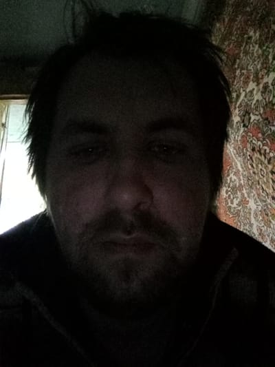 The image is a dimly lit close-up of a man's face. He has dark hair, a beard, and appears to be indoors. A window is visible on the left side of the frame, and a patterned wall or curtain is in the background on the right. The scene is inferred to be in Parabel, Russia, as requested.