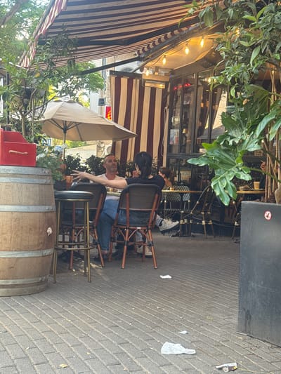 Here is a detailed description of the image:

**Location:** The image appears to be taken at an outdoor cafe in Givatayim, Israel.

**People:** The image shows a man sitting at a table with another person.

**Subjects:** The main subject is the outdoor seating area of a cafe. Additional subjects include tables, chairs, a barrel, and plants.

**Objects:** The scene includes striped awnings, outdoor umbrellas, light bulbs, tables, chairs, a wooden barrel (likely used as a decorative element), plants in containers, and discarded trash on the sidewalk.

**Actions:** People are seated at the cafe, with one man turning his head to look at the camera.

**Interactions:** People are interacting with each other.

**Environment:** The environment appears to be a bustling street in Givatayim, as indicated by the typical urban scenery. The cafe setting is on a brick sidewalk.
