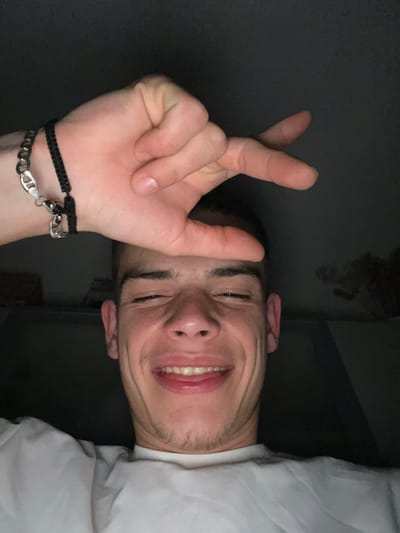 A young man is angled at 90 degrees, so his head is in the right side and his arm is on the left side of the frame. He is wearing a white t-shirt, and two bracelets on his left wrist, one black and one silver. He is making a gesture with his left hand. The location cannot be confirmed and is likely not in Sopot, Bulgaria.