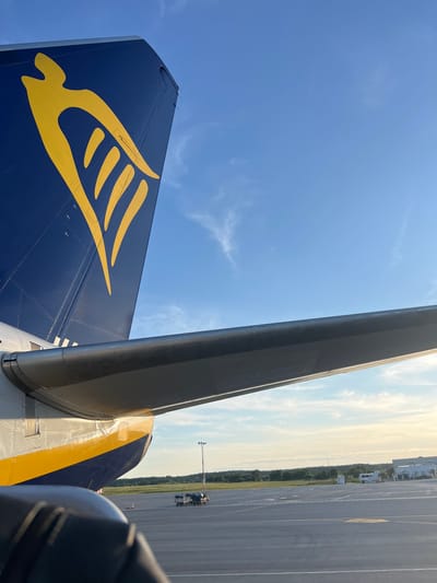 The image shows the tail of a Ryanair airplane with its distinctive yellow logo against a blue sky. A portion of the airplane's wing is visible in the foreground. The scene is set on an airport tarmac, with baggage carts and airport buildings visible in the background. The presence of these elements suggests the image was taken at an airport. The question specifies the location of Karmėlava, Lithuania. Karmėlava is known for Kaunas Airport (KUN), which serves the city of Kaunas. Therefore, the image is likely taken at Kaunas Airport in Karmėlava, Lithuania.