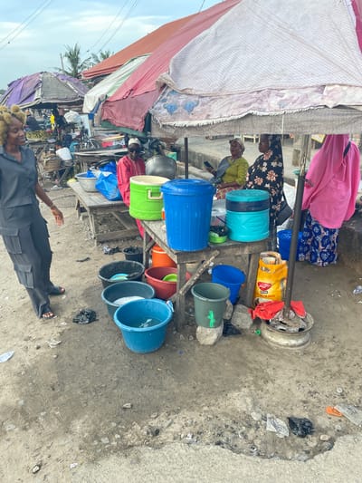 The scene depicts a street in Badagry, Nigeria. A market stall displays various colorful buckets, plastic containers, and cookware. Two women are standing behind the stall, one in a green top and another in a floral dress. A bag of "PRIMA FOODS MOLINA" is visible among the goods. A man in a dark suit and sandals stands in the foreground. The ground is cracked and littered with debris. Overhead, a makeshift awning provides shade. In the background, there are other stalls and pedestrian activity.