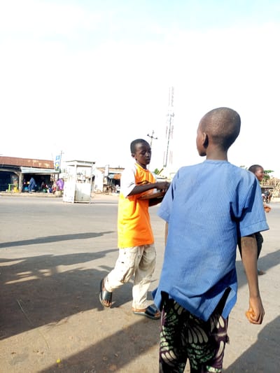 In Tumfure, Nigeria, three young boys are seen standing on a paved road in what appears to be a commercial area. The boy in the foreground, facing away from the camera, wears a blue striped shirt and patterned trousers. To his left, another boy in an orange t-shirt and light-colored pants holds a tray. A third boy is partially visible to the right, smiling. Buildings line the street, and a mobile tower is visible in the background. The sky is bright and overcast.