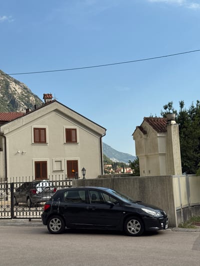 The image captures a street scene in Donji Orahovac, Montenegro. A black hatchback car is parked on the paved road in the foreground, facing right. Behind it, a black SUV is visible through a decorative metal gate, which encloses a property with a light-colored house. The house has dark brown shutters on its windows. To the right of the house, a tall, pale structure with a tiled roof stands, and further in the background, a dense line of green trees is present. Mountains with sparse vegetation rise in the distance under a clear blue sky. The scene appears to be a typical residential area.