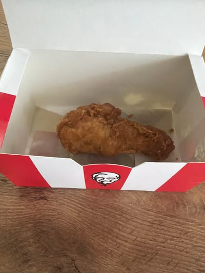 A single piece of fried chicken is centered within an open white and red striped cardboard box. The box is placed on a wooden surface. The chicken appears to be a drumstick or a similarly shaped piece, coated in a golden-brown breading. The box has a logo on its front that is partially visible. The scene is lit from above, casting soft shadows. The background is a plain white interior of the box, with hints of the wooden surface visible around the edges. The image does not contain any contextual information that would allow for its localization to Bila Tserkva, Ukraine, or any other specific city.