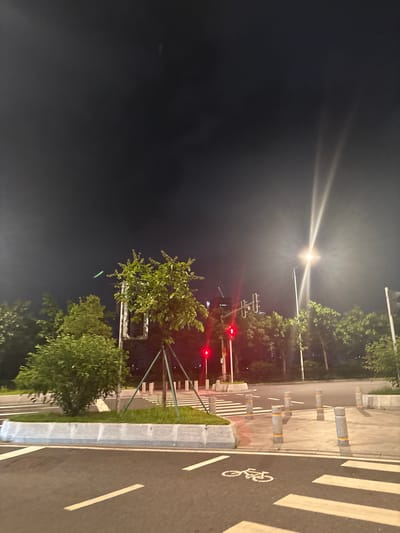 The image depicts an urban street scene at night in Tianhe District, China. The sky is dark and overcast, with bright light emanating from a street lamp on the right side of the frame, creating strong light streaks. A traffic light showing red is visible in the middle ground, along with other traffic lights further in the background. Trees with green foliage are scattered throughout the scene. In the foreground, a road with white lane markings and a bicycle symbol is visible, along with a pedestrian crossing marked by white stripes. Several bollards with yellow bands line the edge of the crossing and sidewalk. A concrete barrier separates the road from a raised grassy area with a small tree supported by stakes. The overall atmosphere is nocturnal and urban, with artificial lighting illuminating the street and surrounding elements.