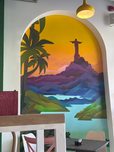 The image shows a mural on a wall, depicting a stylized landscape. In the foreground, a wooden table with an ashtray and a chair are visible. The mural itself features a silhouette of Christ the Redeemer statue atop a mountain range, with a palm tree and a vibrant sunset in the background. The scene is illuminated by a yellow pendant light. The mural is framed by an arched opening. The presence of tables and chairs suggests this is an indoor dining or seating area, possibly a restaurant or cafe. There is no visible indication of the location being in Podgorica, Montenegro.