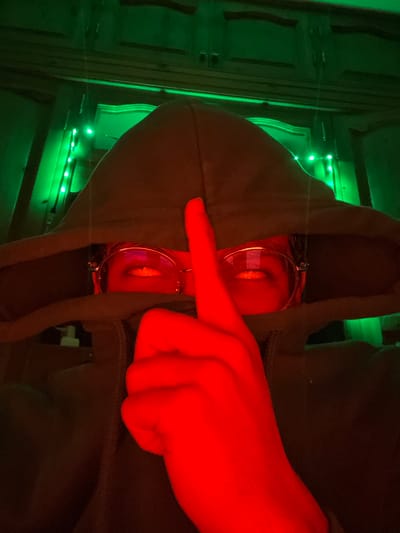 An upside-down image features a person wearing a green hoodie. The hood is pulled up and obscuring the face, but two eyes are visible, magnified and distorted behind glasses. A hand, illuminated in red light, is positioned in front of the hood, with the index finger extended across the hood's edge. Green lights twinkle in the background near a wall or door. The overall aesthetic is dimly lit and somewhat mysterious.