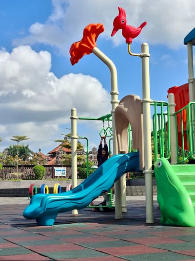 In Gianyar, Indonesia, a brightly colored playground sits under a partly cloudy blue sky. The playground features a blue slide with a frog-shaped base, and a green slide with a frog decoration at its side. In the background, a traditional Balinese temple with intricate architecture and red tile roofs is visible. A child is seen hanging from monkey bars on the playground, their arms extended. The playground is situated on a patterned rubber mat surface.