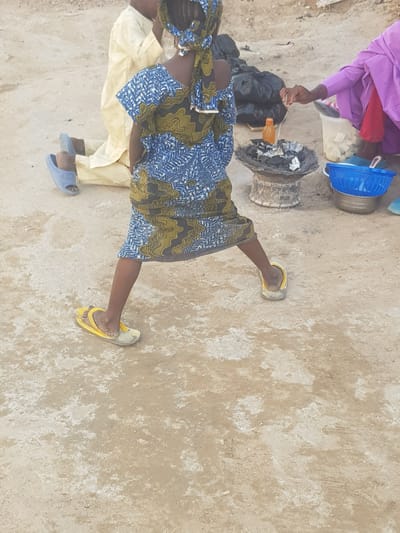 The image depicts two individuals in an outdoor setting in Dabawa, Nigeria. The environment consists of a beige, textured ground. One person wears a blue and green patterned dress and yellow sandals. Another person is wearing a purple top, red pants, and is standing near a charcoal stove. There is a blue basket nearby, as well as a white container, and a black bag next to the stove. One of the individuals seems to be using a thin string to prepare a cooking ingredient above a container of black charcoal and coals. The third person is wearing a cream outfit with blue sandals.