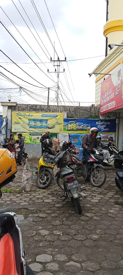 The scene is a street in Cangkuang, Indonesia (latitude: -7.042, longitude: 107.554) with several motorcycles parked on the side of the road. There are several people in the scene, some standing, some sitting, and some walking. Some of the people are sitting on motorcycles, while others are standing next to them. There is a large building in the background, with several signs on it. The signs are in Indonesian and appear to be for a food store. The street is made of cobblestones and there are power lines overhead. There is a large yellow motorcycle in the foreground, a yellow scooter motorcycle, and a black motorcycle. The motorcycles have license plates that are visible. One license plate says "2669 ZTL," while another says "5000 GX." There are a few other motorcycles parked in the background. There are some bags on the ground, and the ground is dirty.