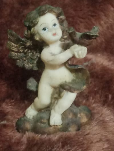 An image of a small, porcelain figurine of a cherub is presented. The figurine is positioned on a dark, fuzzy surface. The cherub has light skin, brown hair, and blue eyes. It is depicted with small, feathered wings and is wearing a draped garment that is green and gold. The cherub's left arm is extended, and its hands are held in front of its chest. The figurine appears to be in motion, with its legs bent as if it were dancing or running. The background is a deep red, textured fabric. The image does not provide any information or context related to the city of Santa Ana, Venezuela.