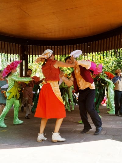 A group of people in colorful costumes perform in a circular gazebo. A woman in a red skirt, patterned top, and headscarf slaps a man's head with a white cloth. The man wears a vest, dress shirt, and dark pants and appears to shield himself. Other performers are dressed as flowers and plants in shades of green and pink. A man with a camera stands to the side, observing the performance. The setting appears to be an outdoor stage.