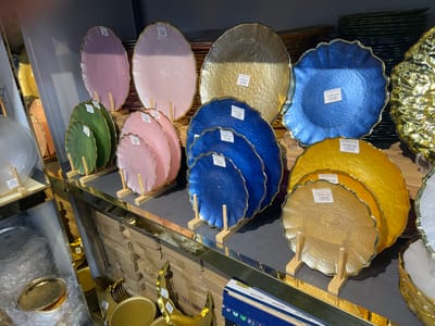 This image displays a retail setting, likely a home goods or kitchenware store in Eshanguzar, Uzbekistan. The foreground and midground are filled with a variety of decorative plates and platters. These items are primarily round with scalloped or irregular edges, and come in a range of colors including pink, green, blue, and gold. Many of the plates are textured and have a glossy, metallic finish.
Several plates are displayed on wooden stands, suggesting they are meant for presentation. Some plates have price tags attached, with text that appears to be in Cyrillic and includes prices in "sum" (the currency of Uzbekistan). For example, visible text includes "55 000 сум", "40 000 сум", and "36 000 сум". Boxes are stacked in the background, some labeled with Cyrillic text and what appear to be numbers like "06c" and "94-25". There is also a box in the lower right corner with text "NEW YORK MOSCOW SOLINGEN".
The lighting suggests an indoor environment, possibly illuminated by artificial store lights. The overall impression is one of an organized display of decorative and functional tableware. No people are visible in the image. The scene focuses solely on the merchandise.