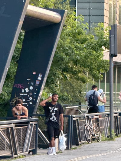 The image captures a street scene in Amsterdam, Netherlands, with a focus on people and urban elements. In the foreground, a dark, angled structure dominates the lower portion of the frame. On this structure, a person wearing a black t-shirt with "TUF AC" printed on it and black shorts is visible, looking upwards and touching their head. Below them, another person with dark hair, wearing a blue bikini top, is also visible, looking towards the camera. The structure itself is adorned with numerous stickers.
In the background, a lush green tree is prominent, with a hint of a building structure behind it. A bicycle is leaning against a metal railing, suggesting a typical urban cycling environment. Several people are seen in the mid-ground, some appearing to be standing or sitting near the bike and railing. One person wears a white cap and a backpack. The overall lighting suggests it is daytime, possibly late afternoon or early evening, with the sun casting shadows and highlighting certain areas. The presence of a bicycle and the architectural style hint at a European urban setting, and specifically Amsterdam. The mood appears casual and relaxed, as if people are enjoying a moment outdoors.