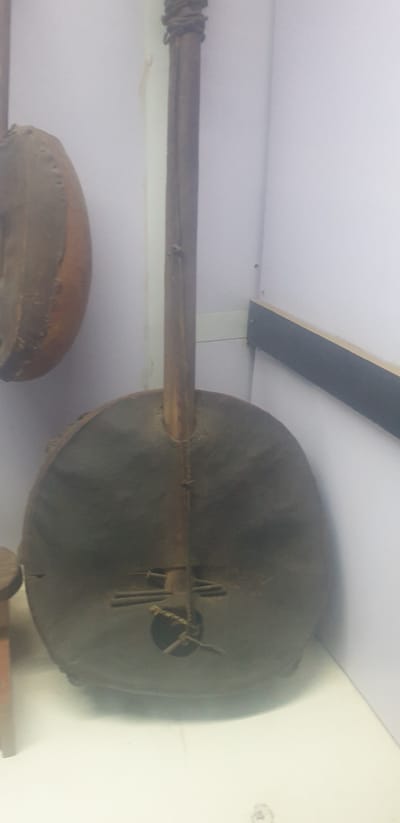 The image depicts a Koora, a West African harp lute, likely located in a museum or exhibition hall in Vom, Nigeria. The Koora has a large round soundbox covered with animal skin. The long neck of the instrument is visible, with a knotted binding near the end. The Koora is mounted on a white wall. A partially visible drum and other unidentified objects are also visible in the lower portion of the frame.