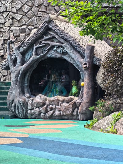 This media file captures an outdoor scene in Sovkhoza imeni Lenina, Russia. The primary subject is a whimsical, grotto-like structure designed to resemble a hollowed-out tree, complete with a thatched roof and a trunk that forms the entrance. Inside this opening, two individuals are visible, appearing to be seated on a bench. Their reflections are visible in the glass pane that encloses them, hinting at this being an exhibit or attraction.
To the right of the entrance, a green, cartoonish lizard-like figure is positioned, and a bright yellow ball hangs from a branch. The structure is built against a backdrop of a large, rough-hewn stone wall. Lush green foliage from an overhanging tree drapes over the thatched roof, suggesting it's daytime and the weather is pleasant, likely spring or summer given the vibrant green leaves.
The ground in front of the structure is a soft, colorful playground surface with abstract patterns of green, blue, and peach. The overall atmosphere is playful and imaginative, suggesting a theme park, children's play area, or a whimsical garden. There is no visible text in the image.
