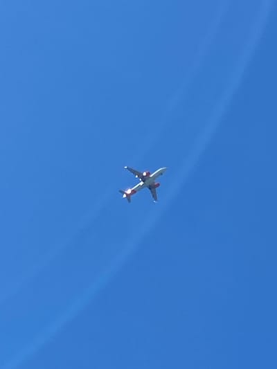 An airplane is captured in mid-flight against a clear blue sky. The aircraft is white with red accents and is positioned horizontally in the frame, viewed from below. It appears to be a commercial jetliner, with its wings extended and its engines visible. The sky is a uniform bright blue, with no clouds. There is a subtle, hazy white streak extending diagonally from the upper right corner of the image towards the center. The image is focused on the airplane, with the background being entirely sky. There are no other objects or people visible in the scene. The location in Prijevor, Montenegro is not discernible from the image itself.