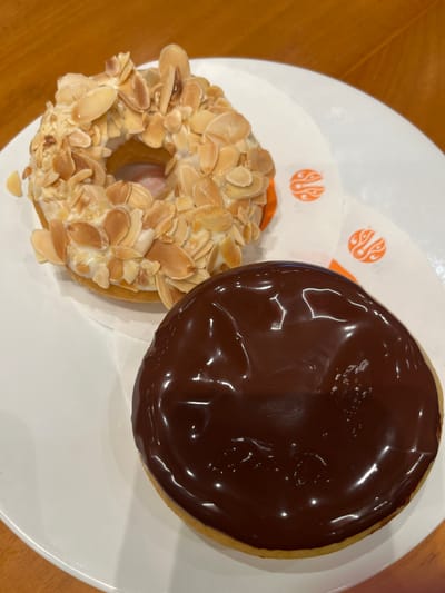 Two donuts are presented on a white plate. The donut in the foreground is covered in dark chocolate icing. The donut in the background is topped with white icing and slivered almonds. The plate is on a wooden table, and the background is slightly blurred. This image was taken in Central Jakarta, Indonesia.