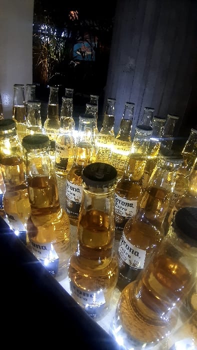A wall of Corona beer bottles, their amber liquid illuminated by subtle lighting. Most bottles are capped and labeled "Corona Extra." The composition creates an abstract, repetitive pattern. Some light shines through the glass, creating a warm glow. At the left of the image is some vegetation with a blue portrait in the background. A dark angled object intrudes from the lower right.