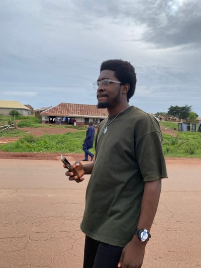 In Durumi, Nigeria, a young Black man wearing glasses and a dark green t-shirt stands on a dirt road. He holds a smartphone in his right hand and a silver necklace with a leaf pendant is visible on his chest. A watch with a black strap is on his left wrist. In the background, several buildings with corrugated metal roofs are situated amidst lush green vegetation. A man in a blue outfit walks away from the camera in the middle ground, and clothes are hanging to dry outside one of the buildings. The sky is overcast with a cloudy sky.