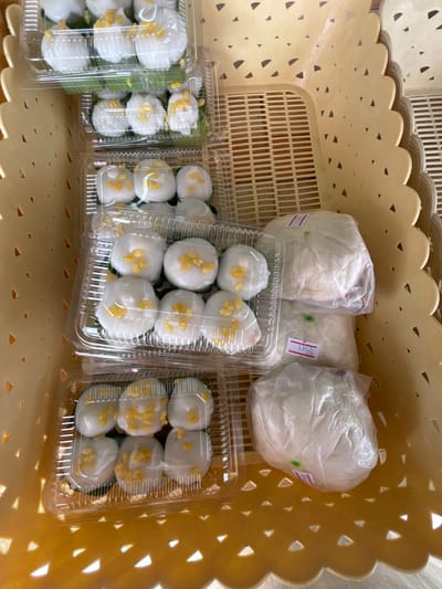 In Rawai, Thailand, a close-up, overhead shot captures a stack of clear plastic containers filled with small, white, dome-shaped desserts topped with yellow kernels, arranged within a yellow woven basket. Three containers are stacked, each containing six desserts. To the right, three larger, white, round food items are wrapped in clear plastic bags and also placed within the basket. One bag has a red-and-white tag with the number "15" visible. The basket has a decorative, scalloped edge with triangular cutouts. The scene appears to be an outdoor market stall or a food vendor's setup.