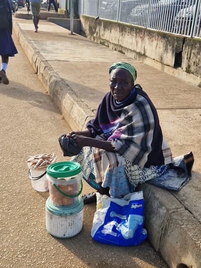 A seated elderly Black woman with a green headscarf and a patterned blanket is on a concrete curb. She is in Kabong, Nigeria. To her left, two buckets are stacked, one containing what appear to be dried shrimp and the other small, fried food items. Next to the buckets is a blue and white plastic bag. To her right, another plastic bag, dark in color, is on the ground. A short wall with some graffiti and a metal fence with cars behind it are in the background.