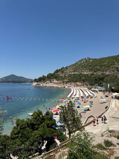 This is a high-angle, wide shot of Kamenovo beach in Montenegro on a clear, sunny day. The beach is packed with white umbrellas, many with people sitting underneath. The sand is a light brown, and the water is a clear, turquoise blue with several people swimming and floating. In the distance, a lush green mountain range slopes down to meet the sea, with some buildings visible nestled into the hillside. There is a pink inflatable water slide in the shallow water near the shore. A concrete pathway with stairs leads down to the beach, and a few people are walking on it. The scene is vibrant and busy, suggesting a popular summer destination.