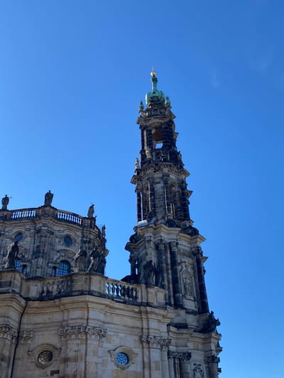 The image is a low-angle shot of the Frauenkirche church in Dresden, Germany, showcasing its Baroque architecture and intricate detailing. The image captures the church's towering spire against a clear blue sky, highlighting the contrast between the stone structure and the sky. The architecture of the church features detailed stone carvings and the spire is topped with a gilded dome.