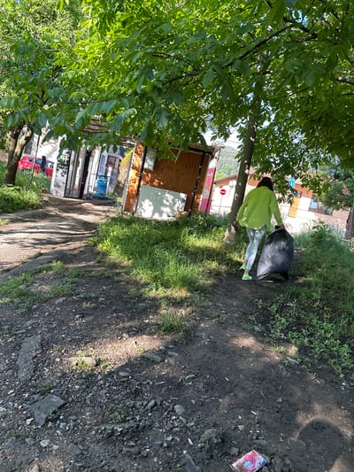 The image captures an outdoor scene in Troyan, Bulgaria, under bright daylight, indicated by strong shadows suggesting late morning or early afternoon. The weather appears sunny and clear.
The primary subject is a person, seen from behind, wearing a bright lime green long-sleeved shirt, light blue jeans, and distinctive bright green sneakers. Their dark hair is pulled back. This individual is holding or moving a large, black plastic trash bag, which they are either collecting or disposing of.
The scene is set on a dirt and gravel path with patches of green grass and weeds. Litter, including a discarded wrapper (partially showing "AIO" or similar text) and cigarette butts, is visible on the ground in the foreground, suggesting a need for cleanup.
To the left, a large tree with abundant green leaves partially frames the shot, its trunk providing a vertical element. In the background, several buildings are visible. Some appear to be simple wooden structures, while others are more substantial with windows, possibly houses or small shops. A red car is parked further down the path. Visible signs include a red and white sign, possibly displaying "DIABLO," and another banner or poster where the word "MONSTER" is discernible. A red circular traffic sign with a white horizontal bar, typically indicating "no entry" or "do not enter," is also present. The overall setting is a semi-rural or peri-urban area, blending natural elements with human habitation.