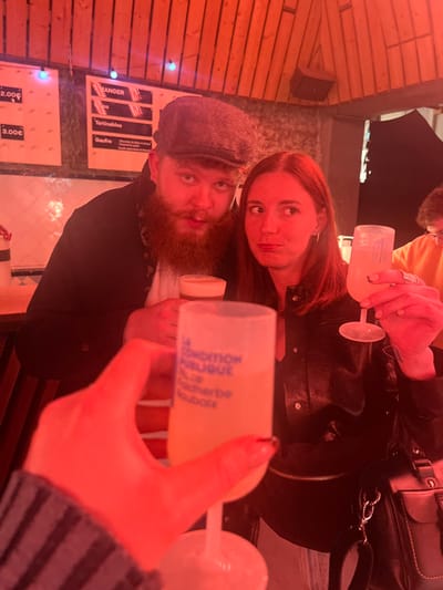 The image depicts three individuals in an indoor, red-lit environment identified as La Condition Publique, Rue Faidherbe, Roubaix, France.

In the immediate foreground, a hand with dark manicured fingernails holds a clear plastic stemmed glass containing a light yellow beverage. Blue text on this glass reads: "LA CONDITION PUBLIQUE R. de Faidherbe Roubaix."

Two central figures are present. On the left, a male with a prominent reddish-brown beard and mustache wears a gray flat cap and a dark jacket over a white shirt. He looks directly at the camera with a slightly open mouth, holding a glass of amber-colored liquid topped with foam. On the right, a female with long, reddish-brown hair, dressed in a black leather-like jacket, looks to her right (viewer's left) with a puckered mouth. She holds a second clear plastic stemmed glass, identical to the foreground glass, containing a light yellow beverage. A black shoulder bag hangs over her left shoulder.

The background features wooden slat paneling on the walls and ceiling. A partial menu board on the left wall displays prices such as "2.00€" and "3.00€", and items including "MANGER" (Eat), "Tartinables", and "Gaufre" (Waffle). Small blue string lights are visible in the upper left background. A blurred partial figure wearing an orange top is visible in the far background on the right. The entire scene is bathed in a dominant red light.
