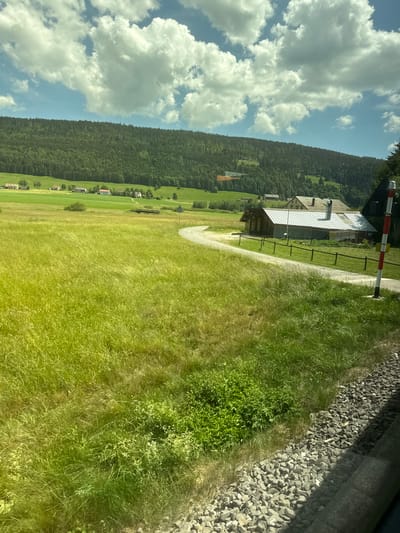This is a landscape photograph, taken vertically, of a rural scene in Le Sentier, Switzerland. The image features a vast expanse of green, grassy fields sloping upwards towards a dense forest that covers a mountainside. The sky is a bright blue, dotted with fluffy white clouds. In the foreground, a gravel track winds through the grass, leading towards a cluster of buildings.
One building, appearing to be a small house or barn with a metal roof, is prominently featured. Adjacent to it is a red and white striped barrier, suggesting a crossing, possibly for a path or a small road. Scattered throughout the lower slopes of the mountain are several small houses with red roofs, creating a picturesque village-like atmosphere.
The time of day appears to be daytime, given the bright sunlight and clear sky. The weather seems pleasant, with the sun casting gentle shadows and highlighting the lush greenery. The overall mood is one of tranquility and natural beauty, characteristic of a Swiss alpine setting. There are no visible people or animals, and no discernible text in the image. The focus is on the serene landscape and the rustic architecture.