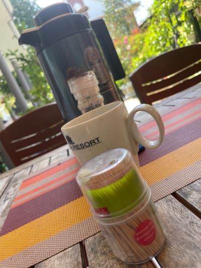 This media file displays a still life arrangement of objects on a wooden table outdoors. The primary subjects are a dark thermos, a white mug, and a clear plastic container filled with toothpicks. The mug has the word "RESORT" printed on its side. The thermos has a circular logo with a number "71" inside. The scene is set on a table covered with a striped placemat in shades of orange, purple, and yellow. In the background, blurred green foliage and hints of pink flowers suggest a garden or natural setting. The lighting indicates it is daytime, and the overall impression is one of casual outdoor dining or relaxation. The location is identified as Phu Thuy Ward, Vietnam. There is no activity or event taking place, but the objects suggest a pause or preparation for a meal or drink. The image is captured from a top-down, angled perspective. There is some text visible on a red sticker on the toothpick container, but it is too blurry to read.