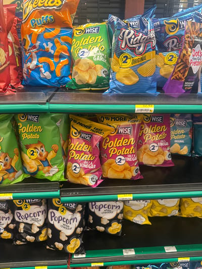 The image shows a close-up, eye-level view of snack food items displayed on store shelves. The shelves are green and black, with white price tags visible. The snacks include bags of Cheetos Puffs, Wise Golden Potato chips in various flavors like Onion & Garlic and Salt & Vinegar, Wise Ridges chips, Dot's Homestyle Pretzels, and Wise Popcorn in White Cheddar flavor. The lighting appears to be artificial store lighting, casting a slight yellow hue on some of the packaging. The background is out of focus, but suggests a retail environment with more products stacked behind. There are no people visible in the image.